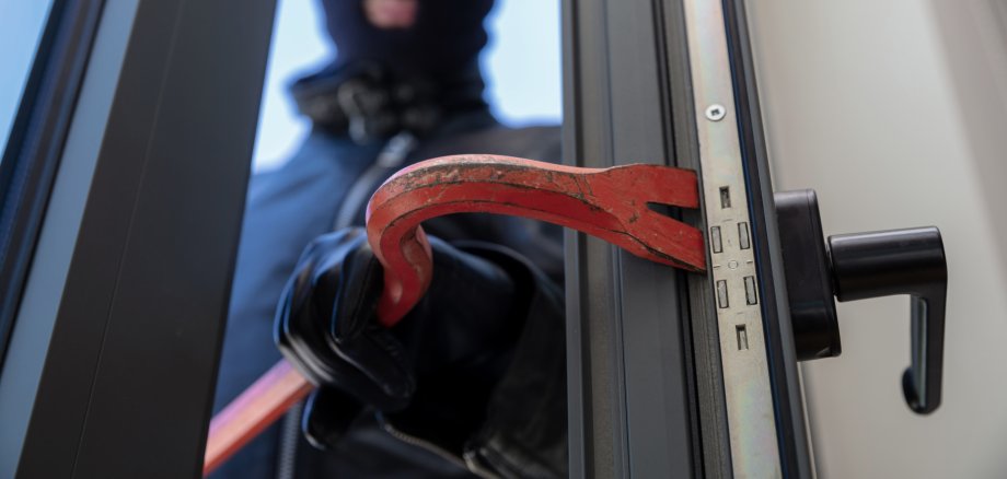 Thief in balaclava holding a crowbar to break a glass window.